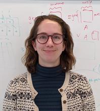 White woman with brown hair and red-framed glasses wearing a knitted cardigan, standing in front of a whiteboard with equations